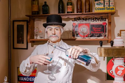 Eine Figur eines Kellners in traditioneller Kleidung, die Getränke in Gläser einschenkt. Im Hintergrund sind Regale mit Flaschen und alten Schildern zu sehen.