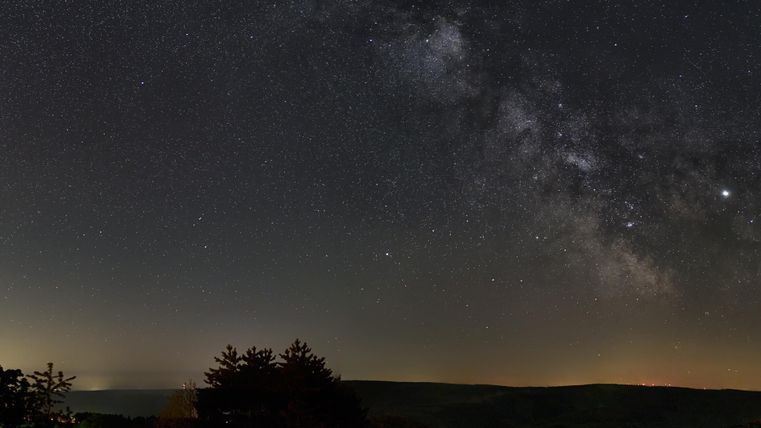 Ein klarer Nachthimmel mit vielen funkelnden Sternen und der Milchstraße. Im Hintergrund sind sanfte Hügel und Bäume zu sehen.
