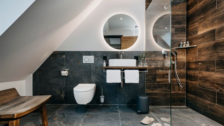 Une salle de bains moderne avec des éléments en bois et en pierre naturelle. Elle dispose d'une douche, d'un lavabo avec un miroir rond et d'une grande fenêtre.