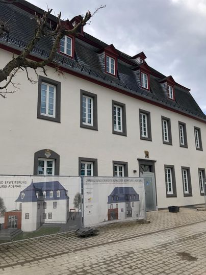 A historic building with a gray roof and white walls. In front of the building, there is a construction fence with a planned project.