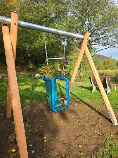 Eine bunte Schaukelschaukel steht in einem grünen Spielplatzbereich. Der Spielplatz ist von Bäumen umgeben und hat eine ruhige Atmosphäre.