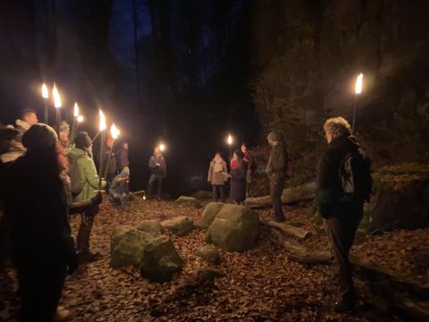Eine Gruppe von Menschen steht in einem dunklen Wald und hält Fackeln. Der Boden ist mit Laub bedeckt und es ist eine mystische Atmosphäre zu spüren.
