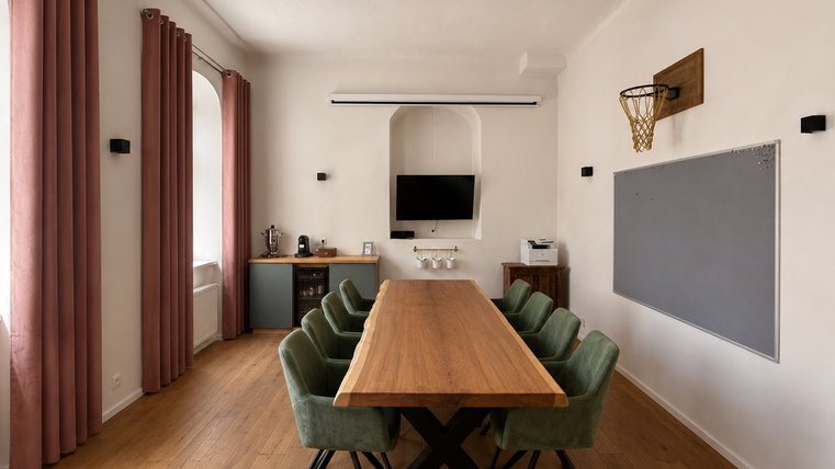 Een moderne vergaderruimte met een grote houten tafel en zes groene stoelen. Aan de muur bevinden zich een televisie, een whiteboard en een basketbalring.