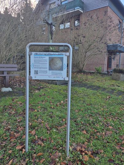 Un panneau d'information se trouve dans un jardin avec des feuilles d'automne. En arrière-plan, un bâtiment et une croix sont visibles.