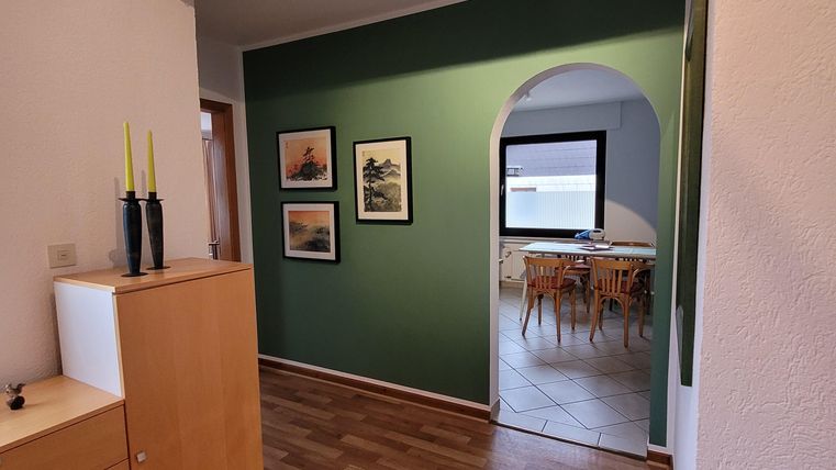 Ein heller Flur mit Holzfußboden und grüner Wand. Durch einen Bogen ist der Essbereich mit einer großen Fensterfront sichtbar.