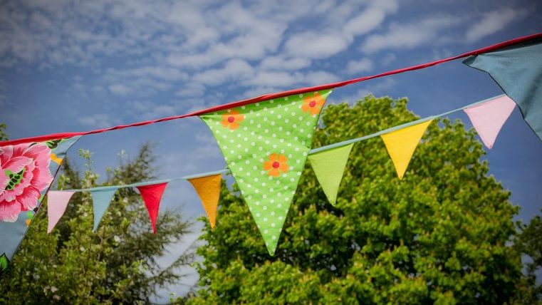 Bunte Wimpel wogen sanft im Wind vor einem strahlend blauen Himmel. Im Hintergrund sind grüne Bäume sichtbar.