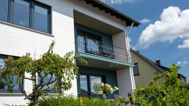 A modern house with a balcony and large windows. Surrounded by green plants and a blue sky.