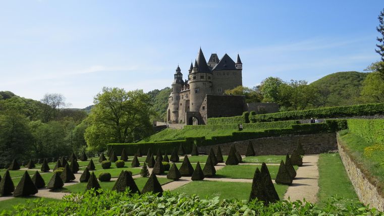 Un château impressionnant entouré de jardins bien entretenus et de collines verdoyantes. Le ciel est clair et bleu.