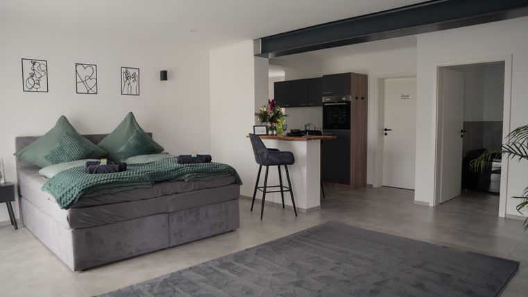 A modern bedroom with a large, comfortable bed and elegant decor. At a small bar, there are chairs, and there is a well-kept, open kitchenette.