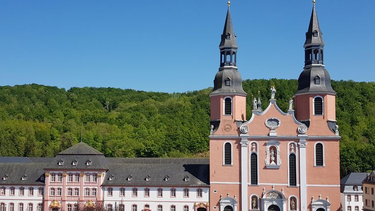 An impressive church with two towers and a red facade. In the background, green hills and a peaceful environment are visible.