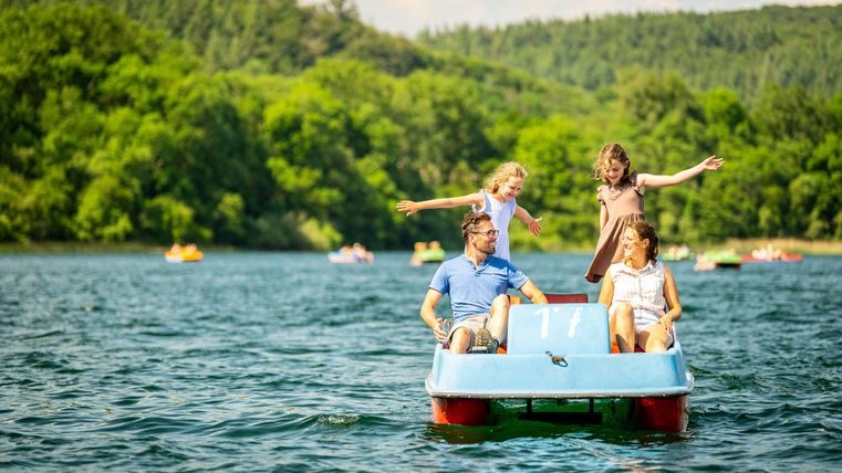 Familie genießt einen Sommertag auf einem Tretboot auf dem Laacher See, umgeben von grünem Wald und klarem Wasser; zwei Kinder stehen fröhlich auf dem Boot, während die Eltern sitzen und lächeln.
