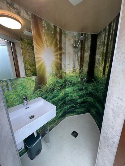 Une douche moderne avec un lavabo et une image de forêt sur le mur. La lumière filtre à travers les arbres et crée une atmosphère apaisante.