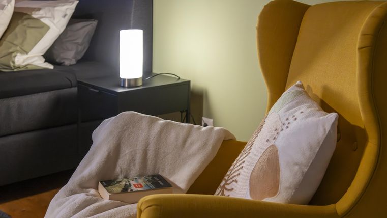 Een gezellige stoel in geel met een deken en een boek erop. Op de achtergrond staat een nachttafeltje met een lamp.