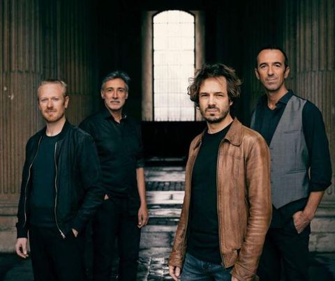 A four-member band stands in front of a window in an impressive architectural space. The members have different outfits and personal styles.