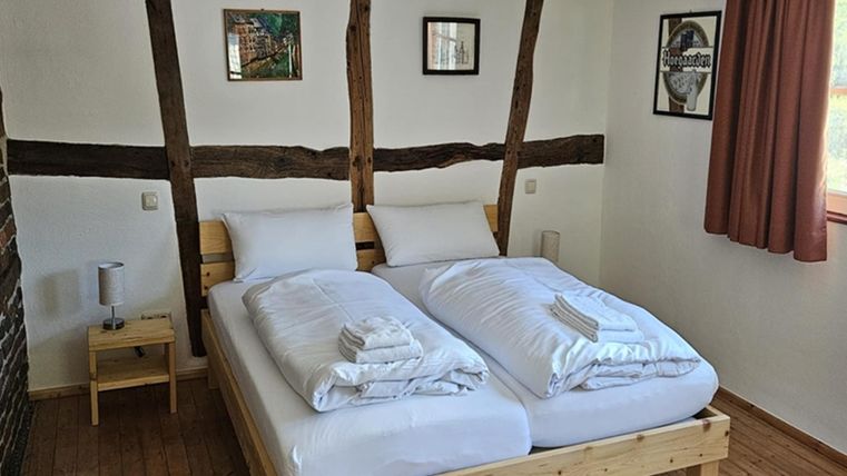 Een gezellige slaapkamer met een tweepersoonsbed en lichte beddengoed. De muren zijn versierd met houten lijsten en er zijn enkele foto's en een raam met een gordijn.