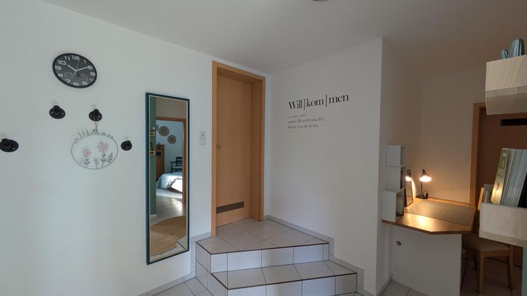 Een lichte entree met een spiegel en een klok aan de muur. De trap leidt naar een andere kamer, en er is een bureau aan de rechterkant.