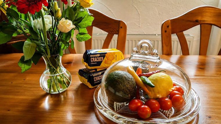 A wooden table with a glass dome under which there are tomatoes, an avocado, and a lemon. Next to it stands a bouquet of flowers and a pack of crackers.