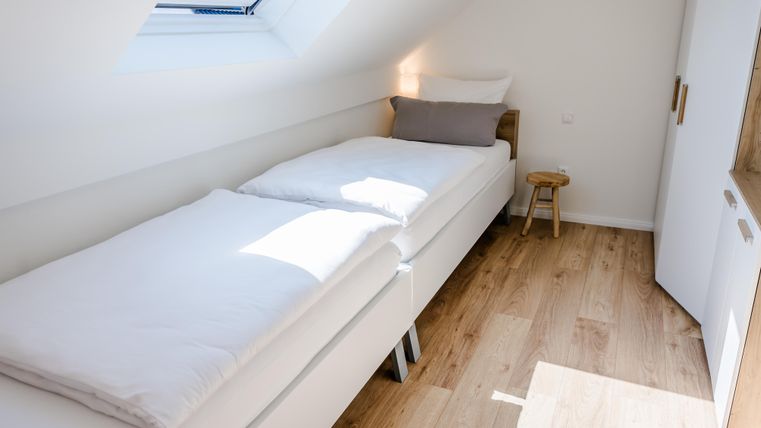 Een lichte slaapkamer met twee bedframes en een kleine kruk. De muren zijn wit en de vloer is van hout.