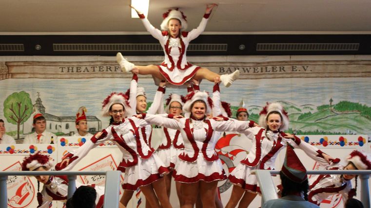 A group of dancers in red and white costumes is performing on a stage. A dancer is standing at the top of a pyramid-like formation.