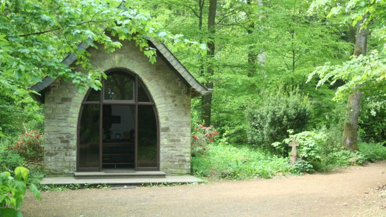 A small chapel in the forest with a stone arch and a cross next to it.