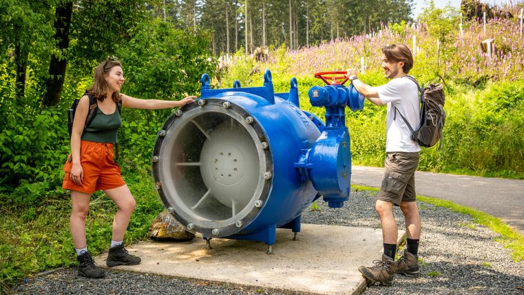 Two people are standing next to a large blue valve in nature. In the background, trees and blooming plants can be seen.
