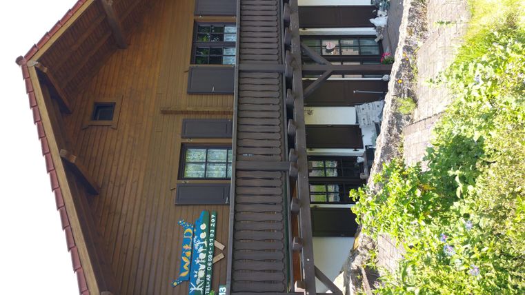 Ein Holzhaus mit mehreren Fenstern und einem Balkon. Es liegt in einer grünen Umgebung.