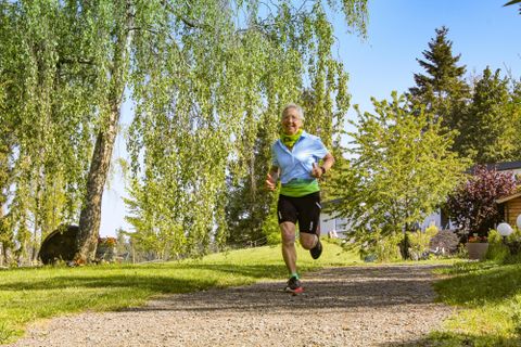Ein älterer Mann joggt auf einem Kiesweg in einem grünen Park. Im Hintergrund sind Bäume und Sträucher zu sehen.