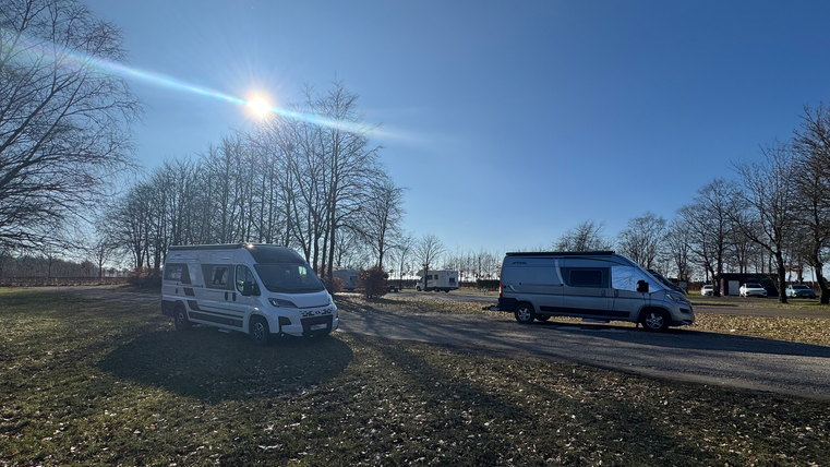Deux camping-cars se trouvent sur une place avec des arbres en arrière-plan. Le ciel est clair et ensoleillé.