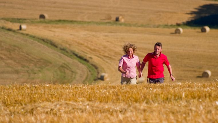 Ein Paar läuft Hand in Hand durch ein goldenes Feld mit Heuballen im Hintergrund.