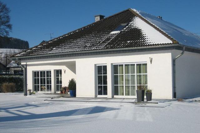 Une maison moderne avec une façade blanche et un jardin recouvert de neige. Le ciel est clair et bleu, et le soleil brille.