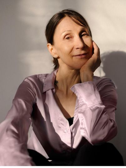 Eine Frau mit schulterlangen Haaren sitzt entspannt, ihr Gesicht zur Seite gedreht. Sie trägt ein helles Hemd und lächelt sanft im warmen Licht.