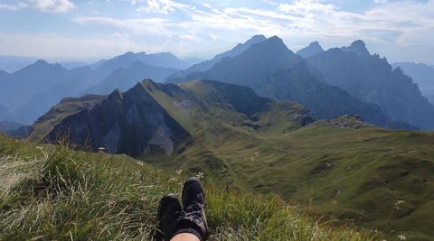 A relaxing view of towering mountains and rolling hills. In the foreground are feet in hiking boots on green grass.