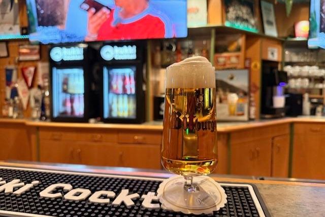 A glass of beer stands on the counter in a bar. In the background, there are vending machines and TVs visible.