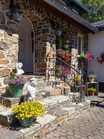 An inviting stone staircase with colorful flower pots. Plants hang on the walls, and the entrance is surrounded by decorative figures.