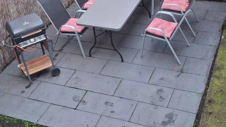 Een gezellige buitenruimte met een tafel en stoelen op een terras. Op de achtergrond staat een barbecue en er zijn grasvelden.