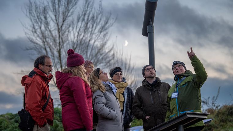 Eine Gruppe von Menschen steht draußen und hört einem Redner zu, der auf einen Punkt zeigt. Der Himmel ist bewölkt und es ist Abenddämmerung.