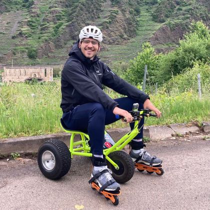 Ein Mann sitzt auf einem kleinen, grünen Fahrzeug mit großen Reifen. Er trägt ein Skateboard-Equipment und lächelt freundlich in die Kamera.