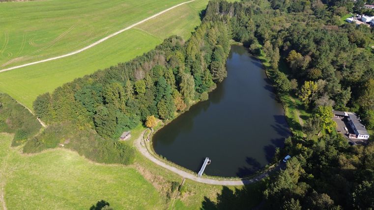 Aerial view of the Gerolstein reservoir, surrounded by dense forest and green meadows. A path runs along the shore, a building is visible.