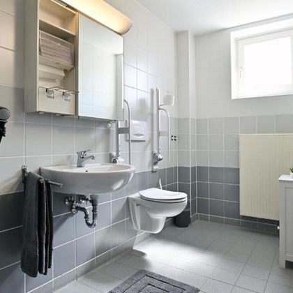 Ein modernes Badezimmer mit grauen Fliesen und hellen Wänden. Es gibt ein Waschbecken, eine Toilette und einen großen Spiegel.