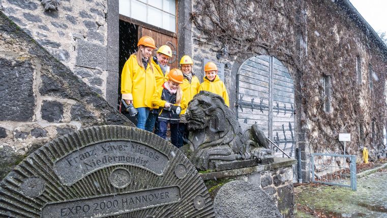 Un groupe de cinq personnes se tient devant un vieux bâtiment couvert de lierre, portant des vestes de pluie jaunes et des casques. À côté d'eux se trouve une pierre avec l'image d'un lion.