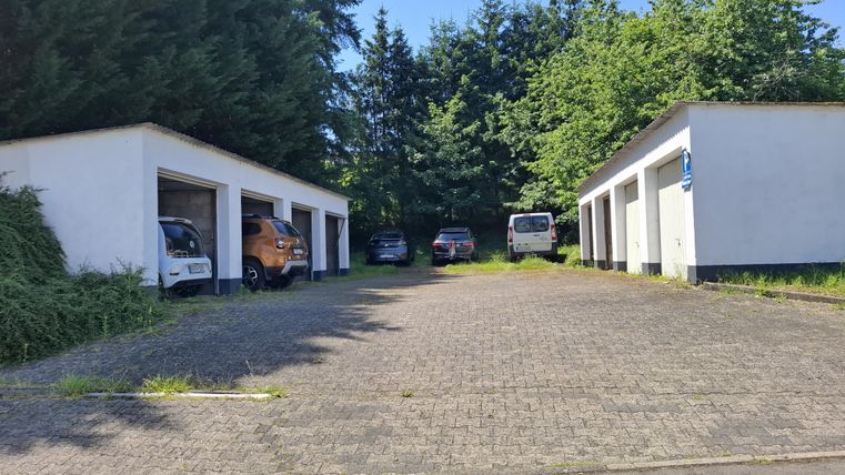Eine Auffahrt mit mehreren Garagen und parkenden Autos. Im Hintergrund sind Bäume und blauer Himmel sichtbar.