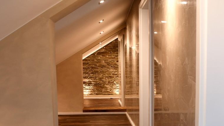 A modern hallway with slanted walls and pleasant lighting. The floor is made of wood and there is a stone wall in the background.
