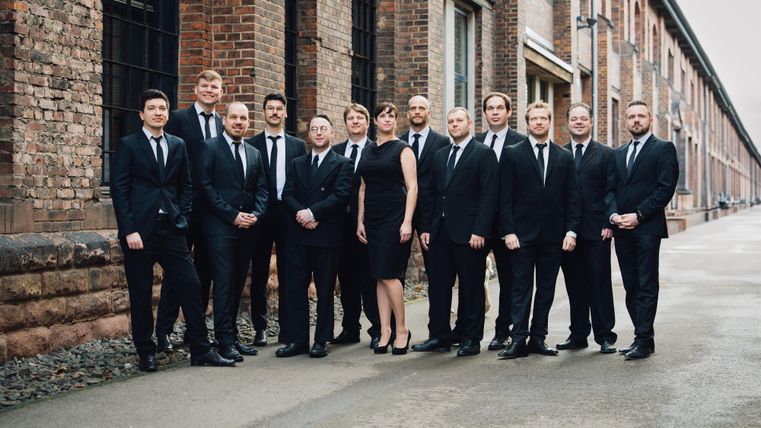 Eine Gruppe von Männern in Anzügen und einer Frau in einem schwarzen Kleid steht vor einem alten Backsteingebäude. Die Atmosphäre wirkt professionell und stilvoll.