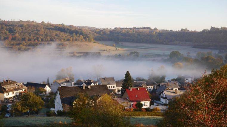 Een rustige landschap met een dorp in de morgenmist. Zachte heuvels en groene weilanden omringen de gebouwen.