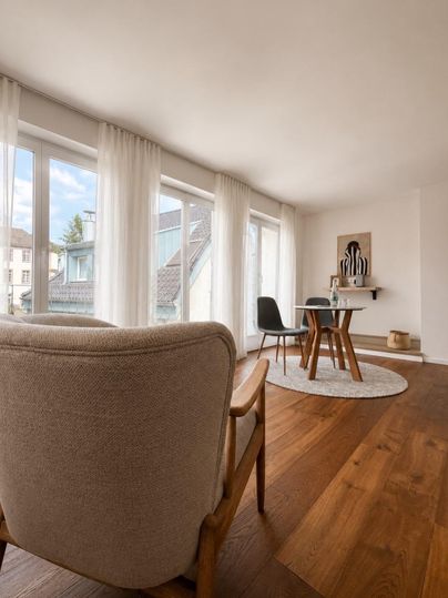 Een lichte kamer met grote ramen en een houten vloer. In het midden staan een tafel en stoelen, aangevuld met een gezellige fauteuil.
