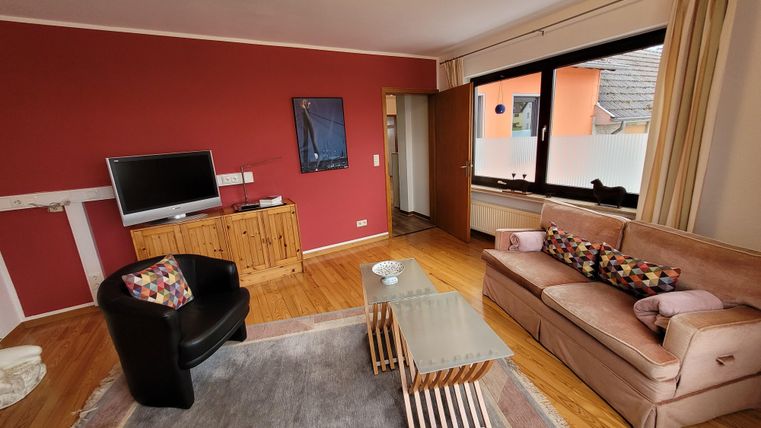 Ein gemütliches Wohnzimmer mit einem roten Akzentwand. Es gibt ein Sofa, einen Sessel und einen Fernseher sowie große Fenster, die Tageslicht hereinlassen.