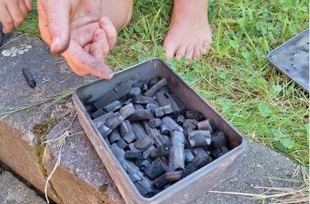 Eine geschlossene Metalldose mit vielen schwarzen Stücken liegt auf dem Boden. Eine Hand zeigt auf die Dose, während barfuß stehende Füße im Hintergrund sichtbar sind.