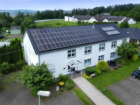 A modern building with solar panels on the roof. The entrance area is surrounded by plants and green spaces.