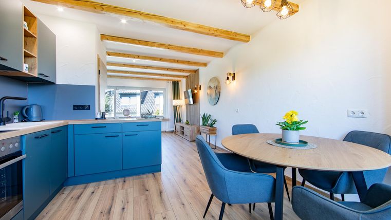 A modern kitchen with blue cabinets and a round wooden table. The room has light walls and exposed wooden ceiling beams.