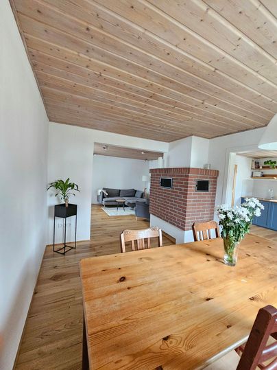 Een modern eetkamer met een grote houten tafel en een gezellige zithoek. Het houten balkenplafond en een tegelkachel creëren een uitnodigende sfeer.